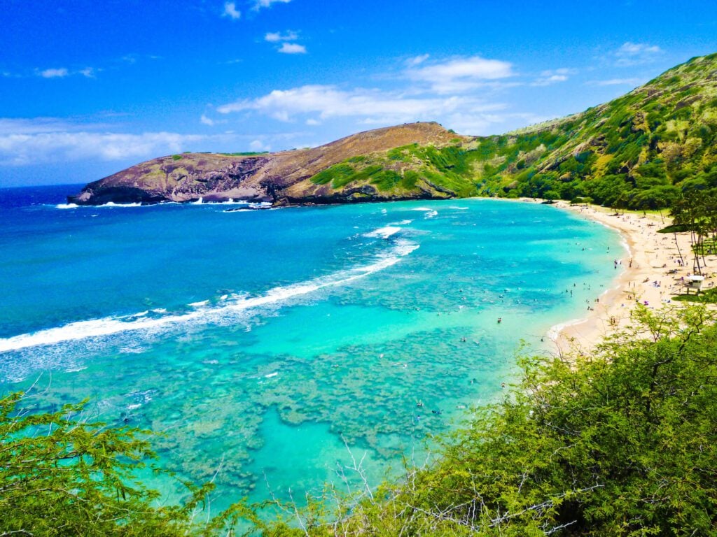 An image of Snorkeling Bay in Oahu, Hawaii featuring a sweeping cove with rolling hills that roll down to aqua colored ocean water containing coral