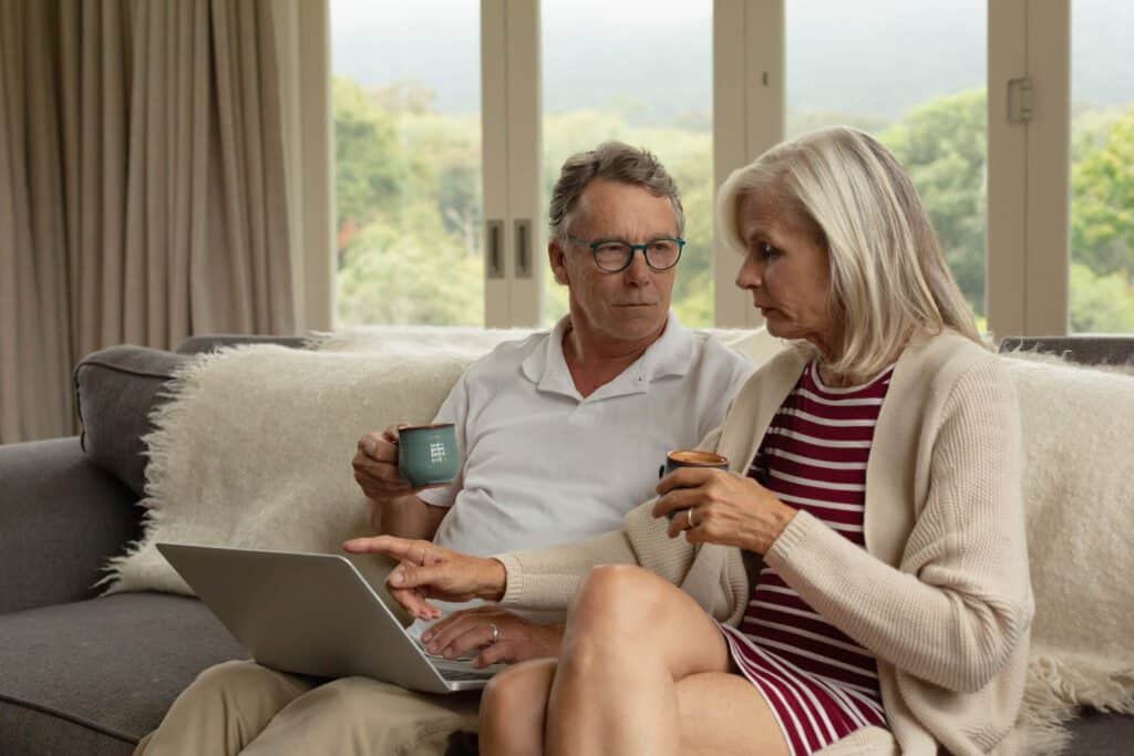 An image of an older couple sitting on a couch drinking coffee as they look at a laptop