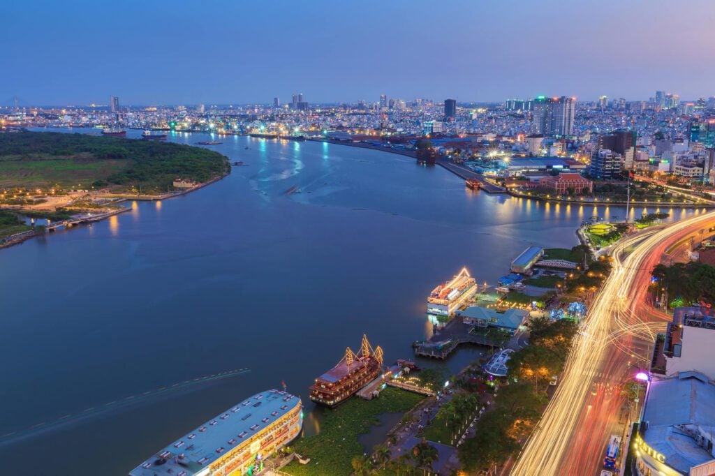 A night time photo of Ho Chi Minh City, featuring a large river and cityscape. There are multiple ferries moored at the riverbank