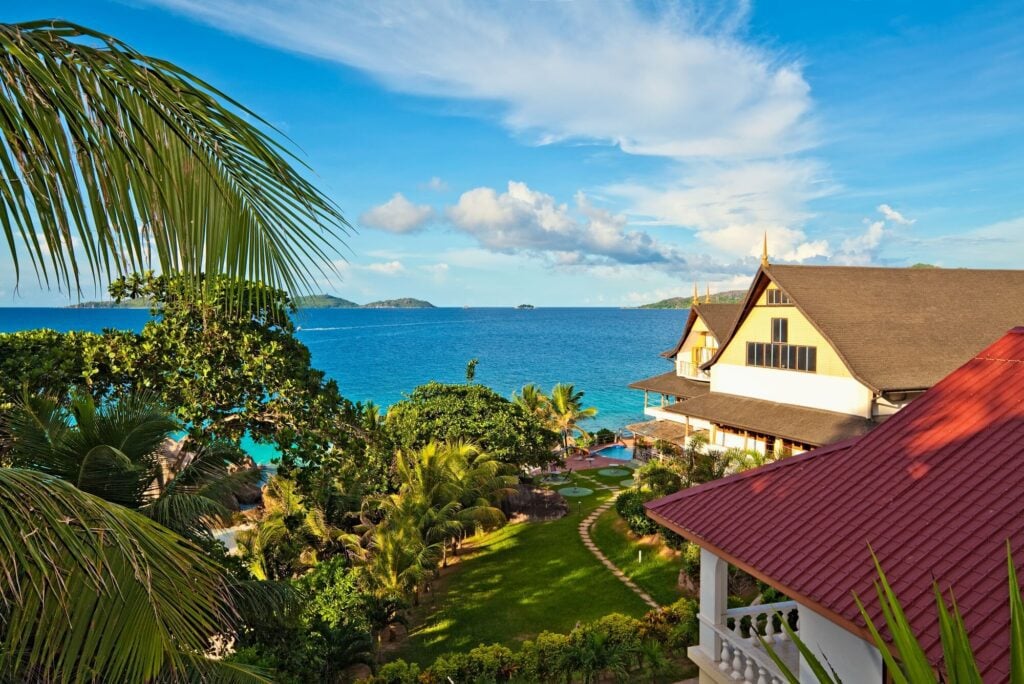 A photo of houses in the Seychelles featuring two large homes, several palm trees, a blue sky and light blue ocean water
