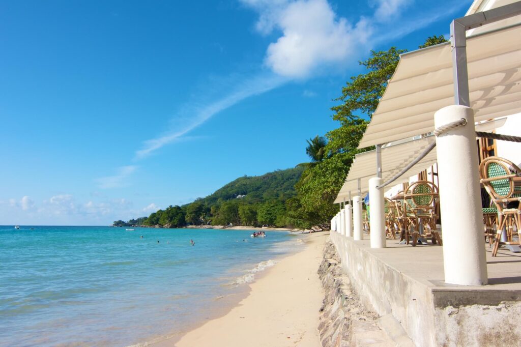 A photo showing a restaurant located on the beach in Seychelles. The small white restaurant is metres from aqua blue water of the carribean