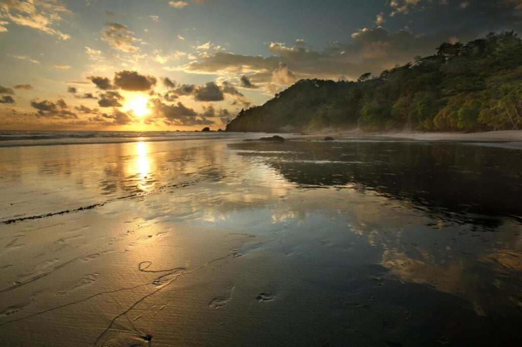 An image of a sunset on a beach in Costa Rica. It shows a wide sandy beach, orange sun hitting the horizon, and an a hill lined with trees to the right