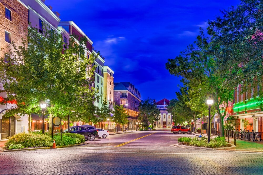 A photo of a Gainsville, Florida street at dusk. It shows a wide street lines with trees and 3 story buildings. The building are very colorful and have shops on the ground floor