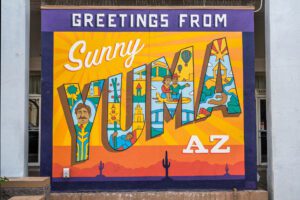 A photo of a colorful sign that says Greetings From Suny Yuma, Arizona. It features bold yellow and orange sun rays, cartoon images of happy people, and a cactus