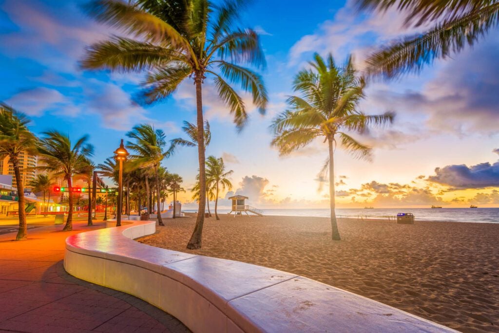 An image of Fort Lauderdale, Florida. It shows a wide sandy beach, several palm trees, a vivid sunset, and a winding footpath. To the left of the image is a number of shops