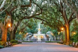 A photo of Forsyth Park in Savannah, Georgia. It shows a wide paved footpath that is lines with huge trees. At the end of a path is a huge water feature with several jets of water